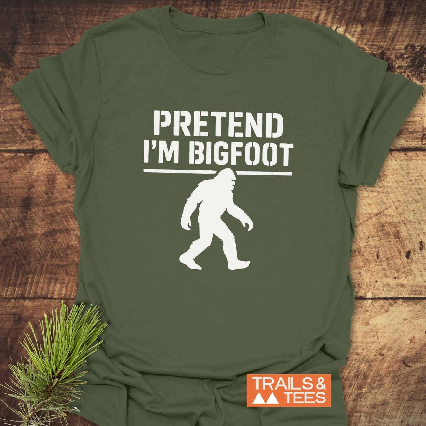 Pretend I'm Bigfoot T-Shirt featuring a Bigfoot silhouette and brand logo, styled with a pine sprig, highlighting outdoor adventure and comfort.