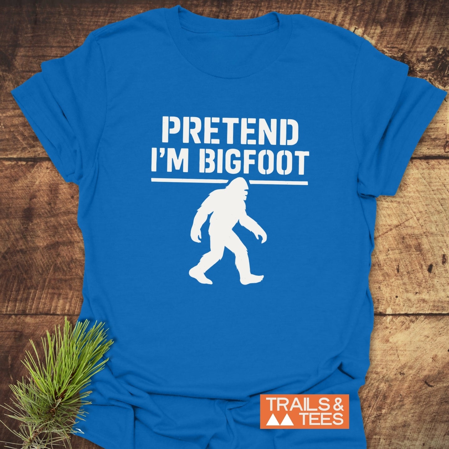 Pretend I'm Bigfoot T-Shirt on wooden surface, featuring a white Bigfoot graphic and playful text, with Trails & Tees label, pine bough accent.