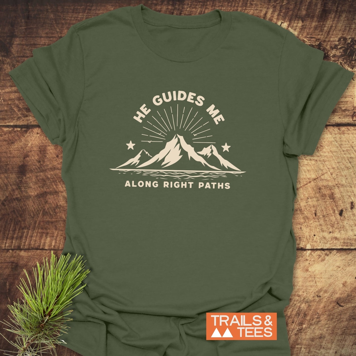 He Guides Me T-Shirt with mountain graphic and inspirational text, laid on rustic wood. Made from soft cotton, ideal for outdoor enthusiasts, durable and pre-shrunk.