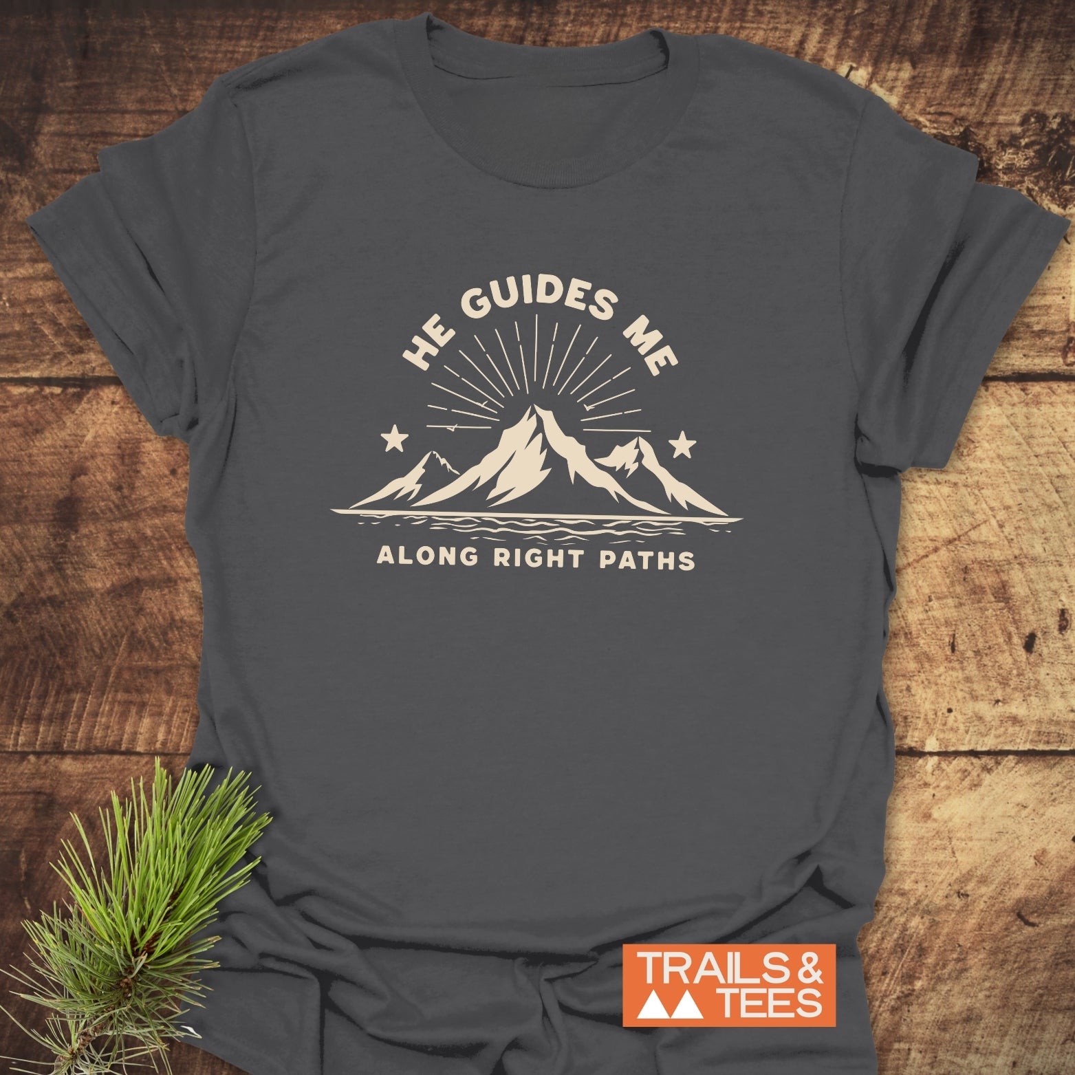 He Guides Me T-Shirt with mountain design and sunbeams, crafted from soft cotton for comfort and durability, featuring the Trails & Tees logo on the corner.