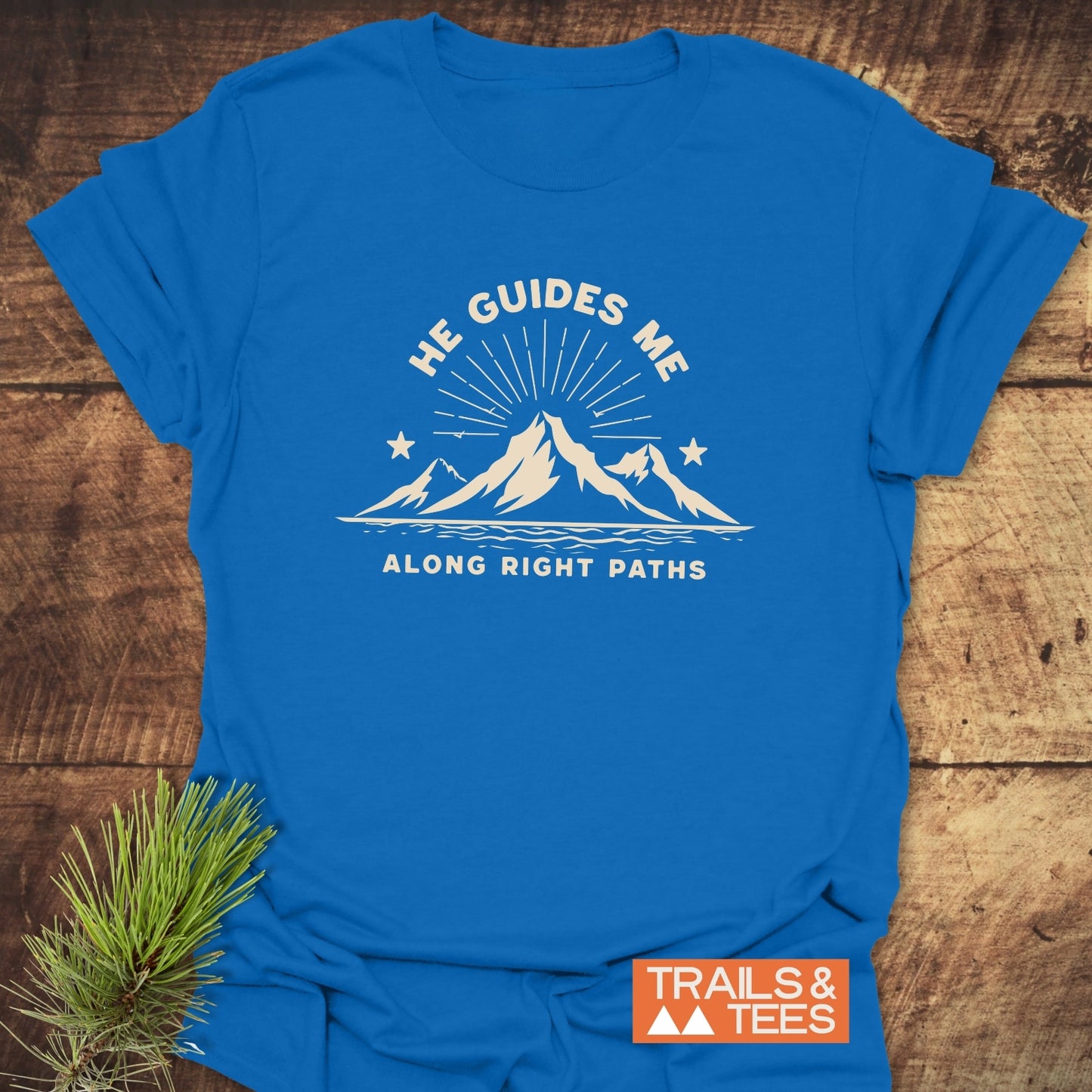 He Guides Me T-Shirt featuring mountain graphic and motivational text, laid on wood. Soft, durable cotton for outdoor enthusiasts, with Trails & Tees logo visible.