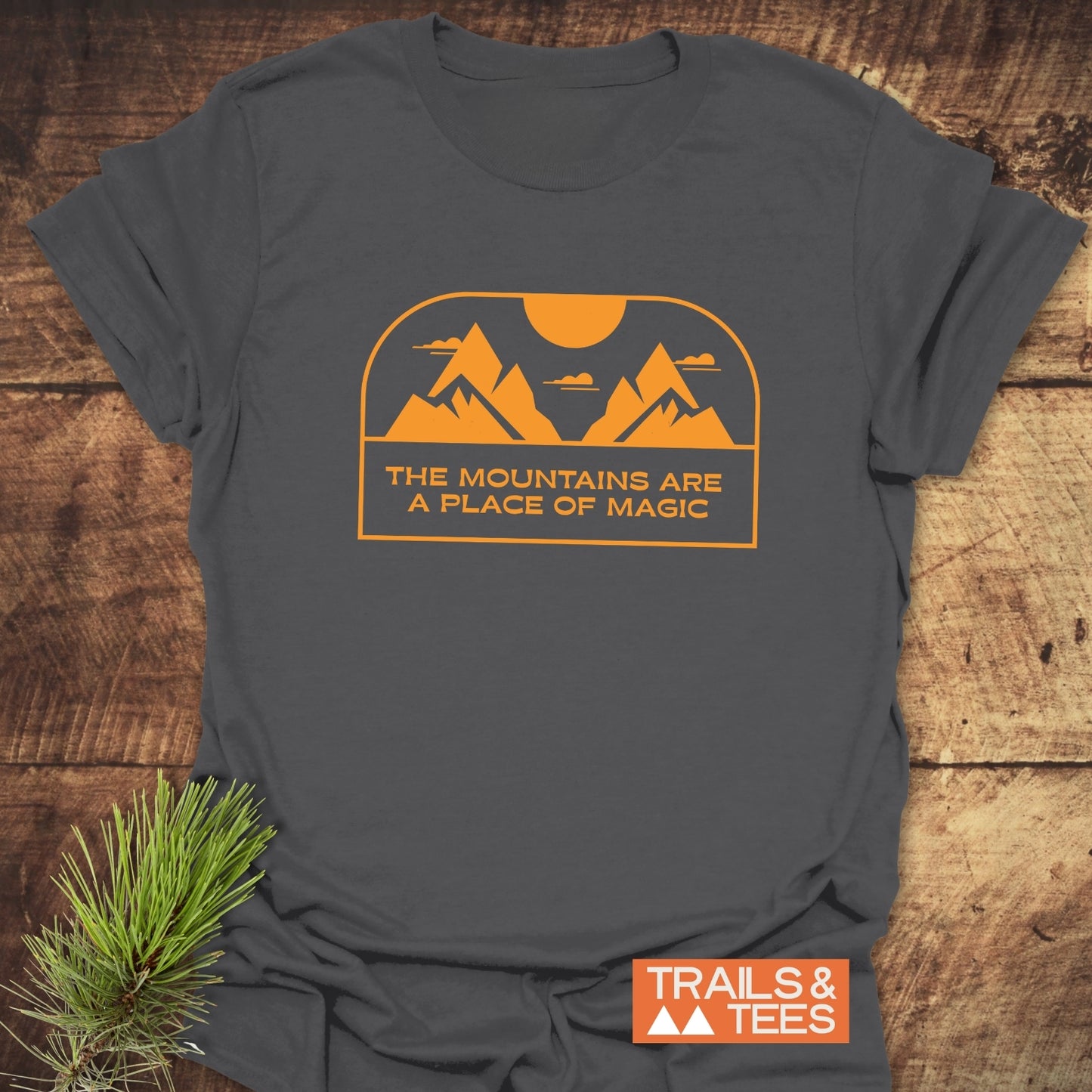 Mountain Magic T-Shirt featuring a mustard/orange mountain graphic and text, laid flat with a pine sprig, showcasing a blend of comfort and outdoor-inspired design.