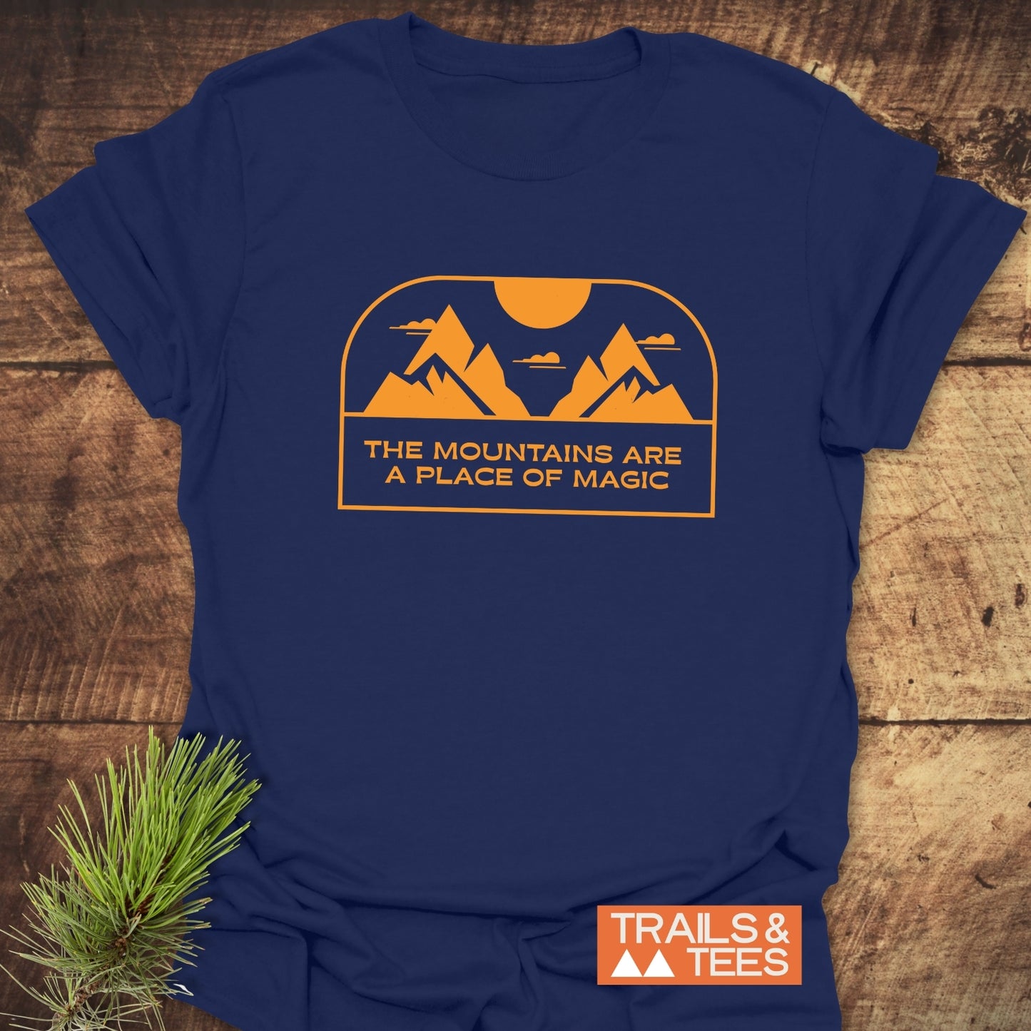 Mountain Magic T-Shirt on wooden surface, featuring orange mountain and sun graphic with text THE MOUNTAINS ARE A PLACE OF MAGIC, beside pine sprig and TRAILS & TEES logo.