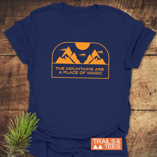 Mountain Magic T-Shirt on wooden surface, featuring orange mountain and sun graphic with text THE MOUNTAINS ARE A PLACE OF MAGIC, beside pine sprig and TRAILS & TEES logo.