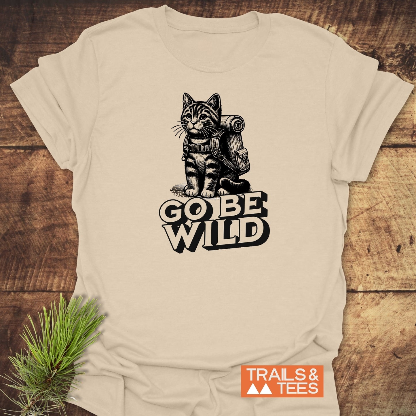 Go Be Wild Cat T-Shirt featuring a cat with a hiker’s backpack and “GO BE WILD” slogan, laid flat with logo “TRAILS & TEES.”