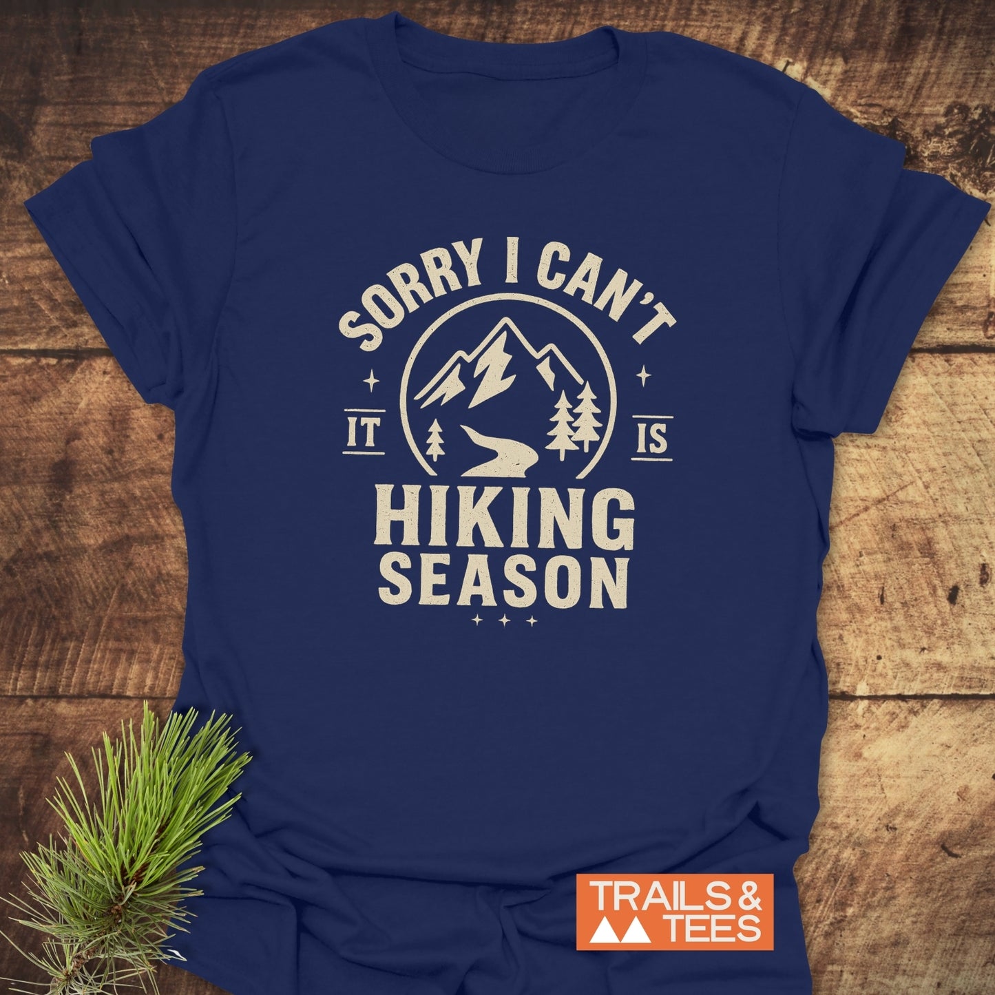 Navy-blue 620 Hiking Season T-Shirt with off-white mountain graphic and text, emphasizing outdoor adventures. Laid flat on wood with pine branch.