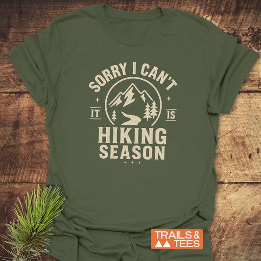 Olive-green 620 Hiking Season T-Shirt with cream graphic of mountains and trees, perfect for outdoor enthusiasts, laid flat with a pine sprig accent.