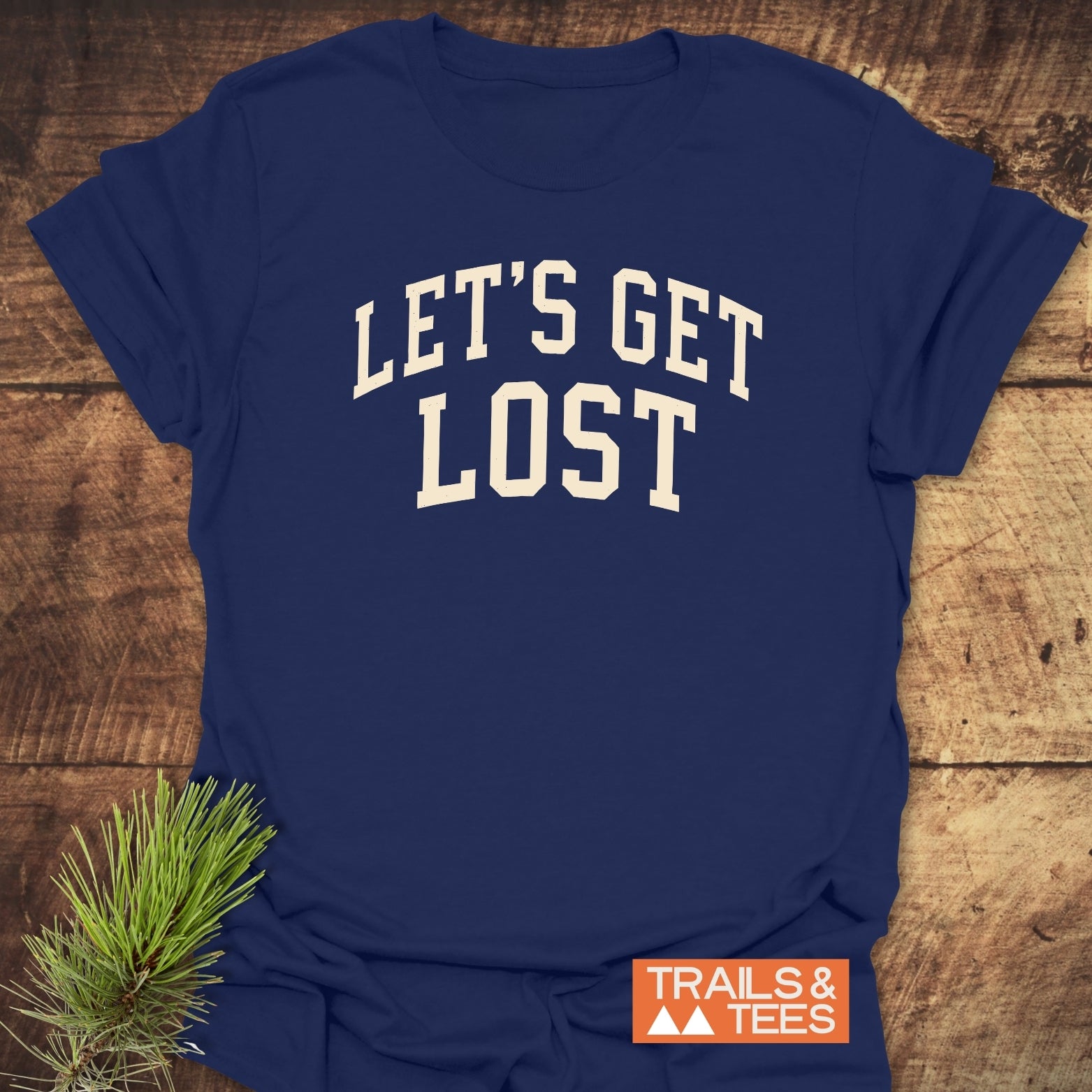 Let's Get Lost T-Shirt flat-lay, showcasing its bold design on soft cotton, with a rustic touch from evergreen needles and Trails & Tees logo.
