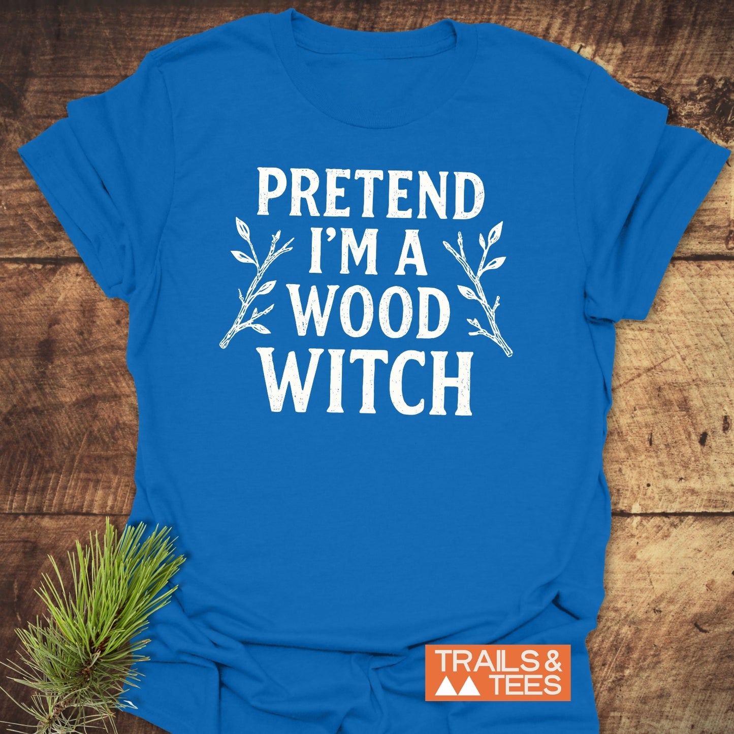 Wood Witch Funny T-Shirt with “PRETEND I’M A WOOD WITCH” slogan and twig graphics, featuring Trails & Tees logo, laid flat on wood surface.
