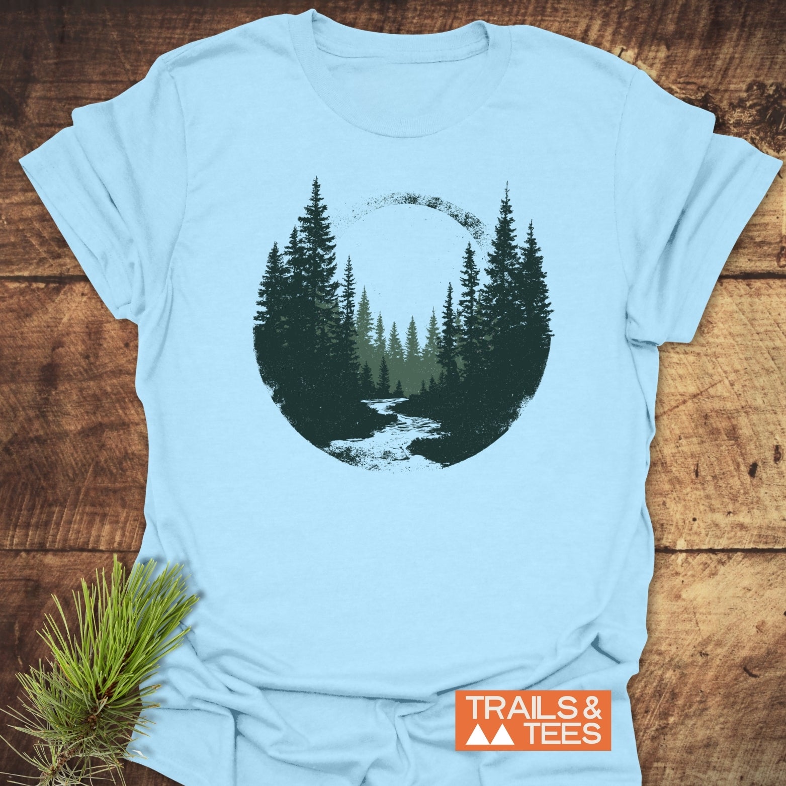Woodland Peace T-Shirt featuring a dark green pine forest scene on light-blue fabric, styled with a pine branch. Comfortable, durable, pre-shrunk cotton for everyday adventures.
