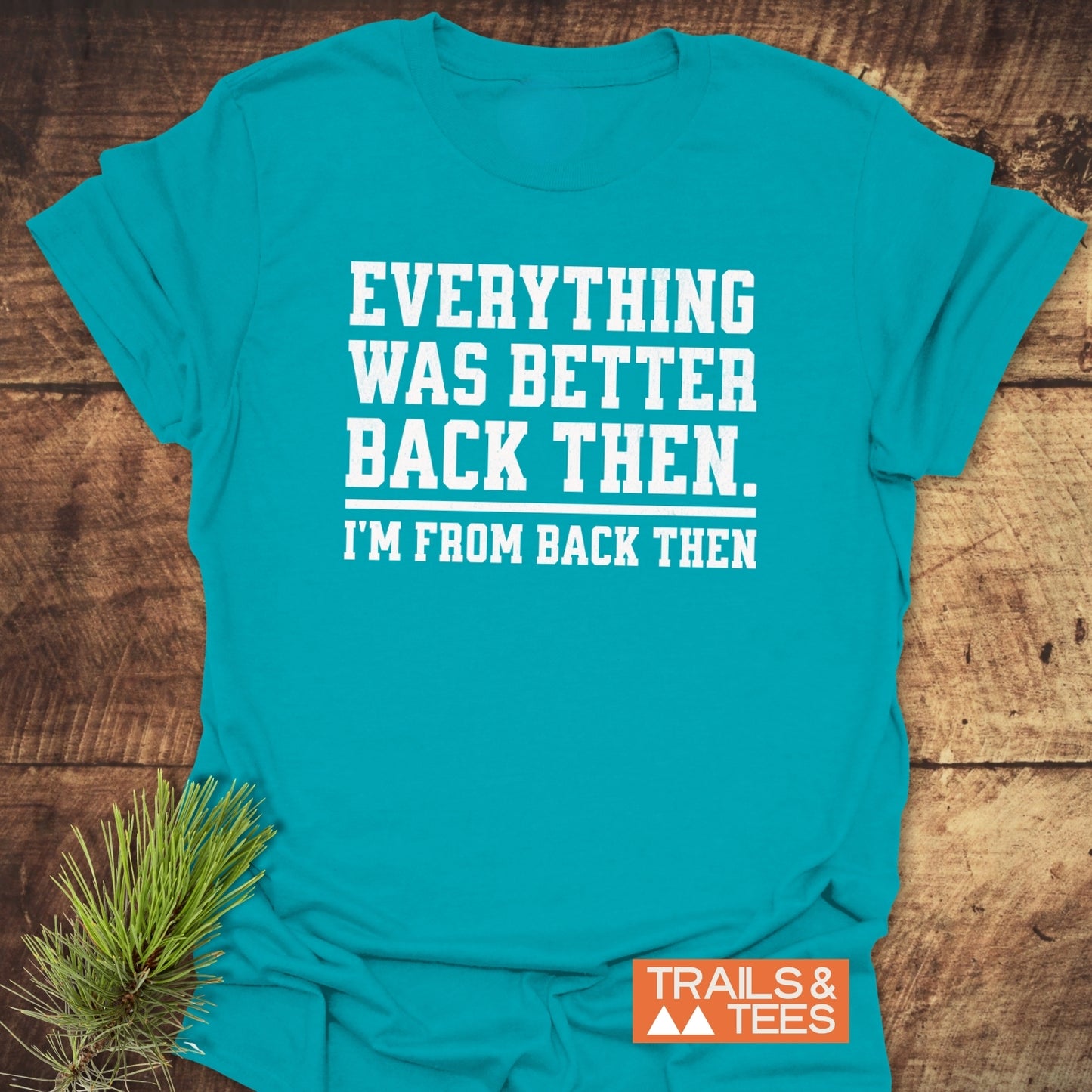 Back Then Funny T-Shirt on wooden surface, featuring white slogan text. Includes Trails & Tees logo and pine prop, highlighting outdoor-inspired, comfortable, durable design.