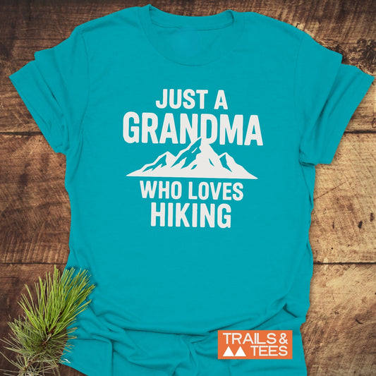 Grandma Loves Hiking T-Shirt featuring a white mountain graphic, emphasizing trail-tested comfort and durable design, laid on a wooden surface with a pine branch accent.
