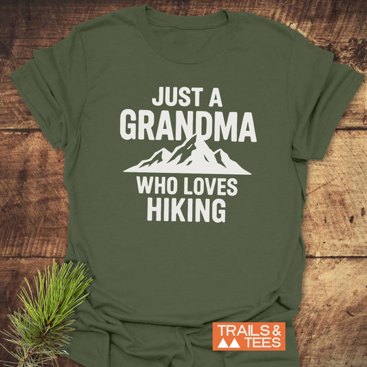 Olive-green Grandma Loves Hiking T-Shirt on a wooden surface, featuring bold white text and mountain silhouette. Includes Trails & Tees logo.