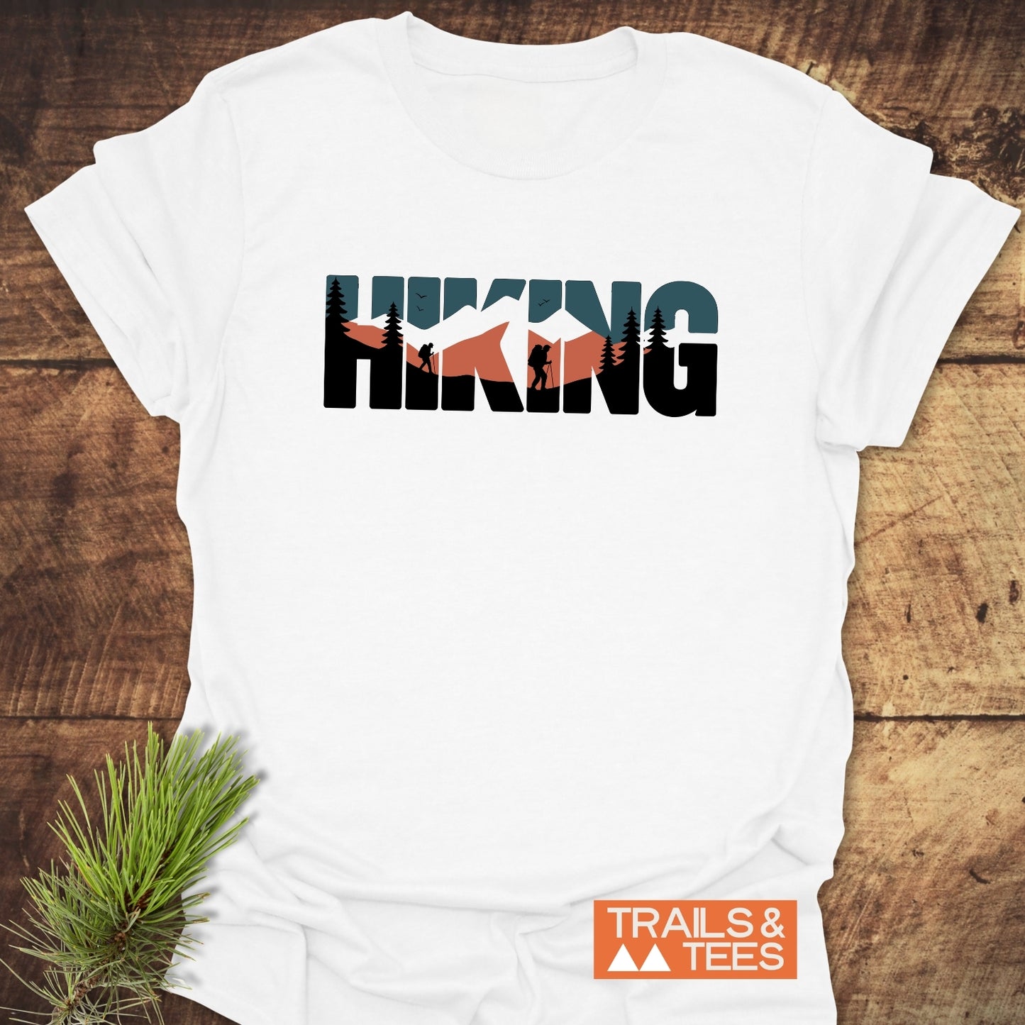 Hiking Retro T-Shirt featuring mountain and forest scenes on letters, laid on wood. Showcases “TRAILS & TEES” logo, crafted for comfort and adventure.
