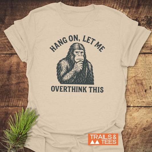 Bigfoot Overthinking T-Shirt featuring a pensive sasquatch print, “HANG ON, LET ME OVERTHINK THIS,” on soft, lightweight cotton, laid on wooden surface.
