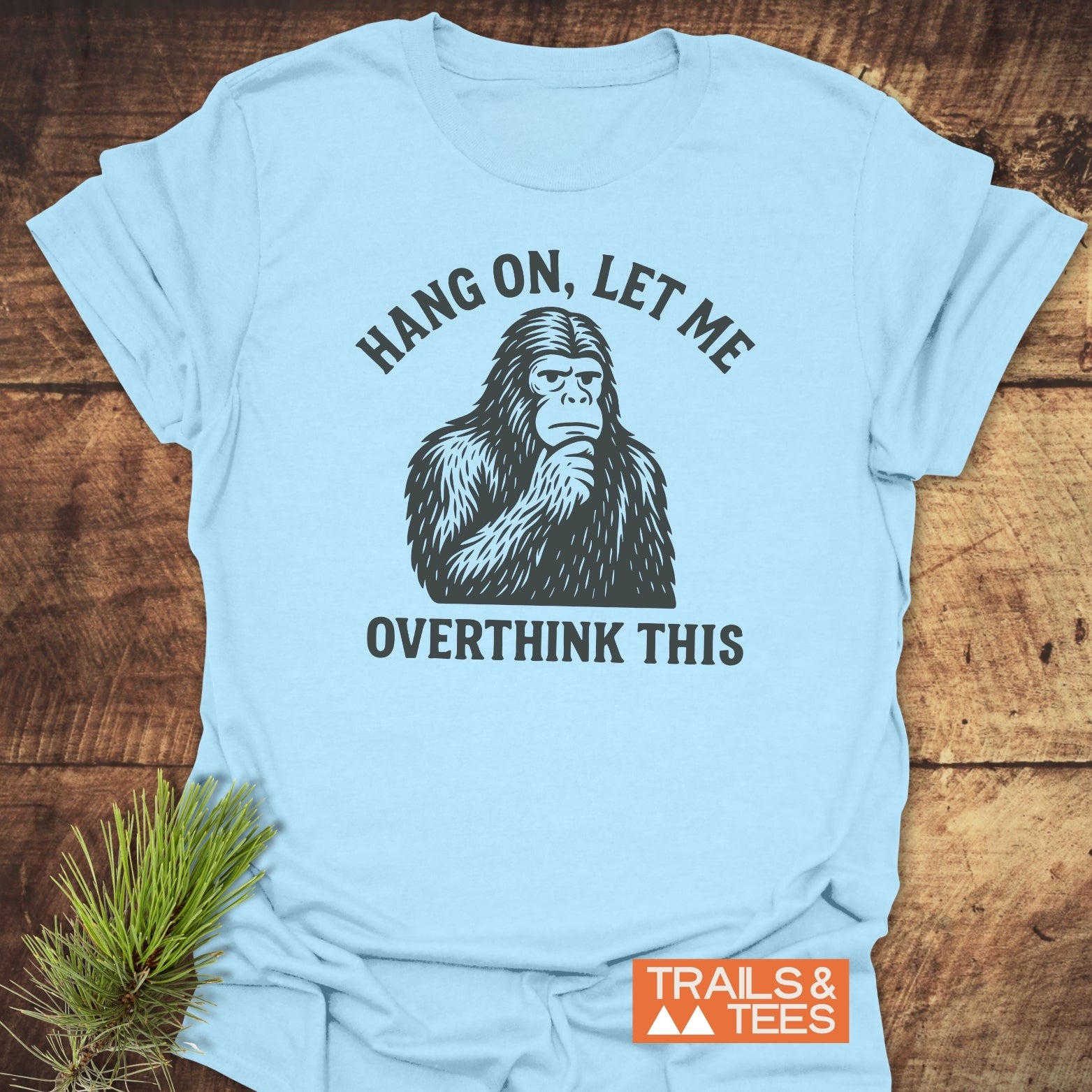 Bigfoot Overthinking T-Shirt on wooden surface, featuring a pensive Bigfoot figure with “HANG ON, LET ME OVERTHINK THIS.” Includes “TRAILS & TEES” logo and pine sprig.