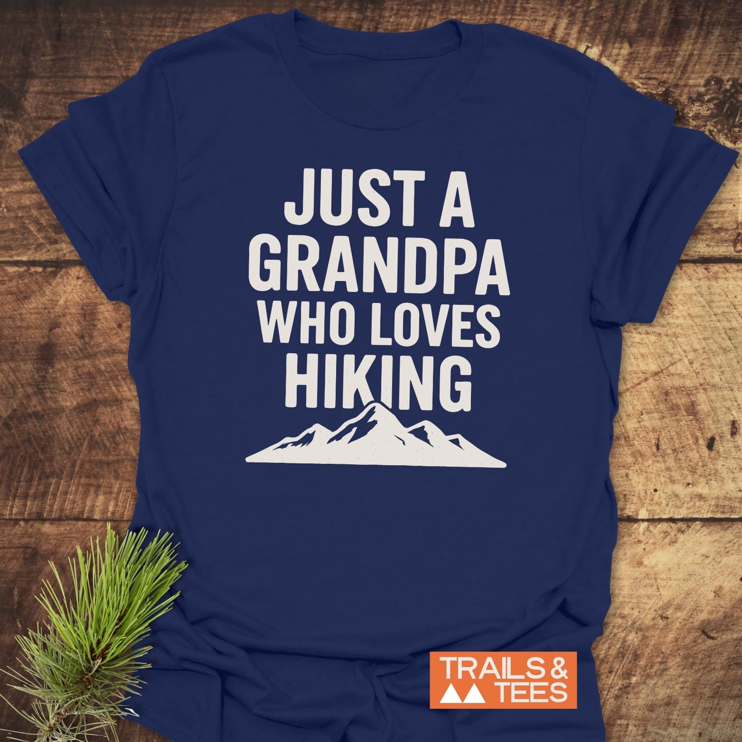 Navy-blue Hiking Grandpa T-Shirt with bold white text and mountain silhouette, laid on wood. Features soft, durable cotton with logo TRAILS & TEES.