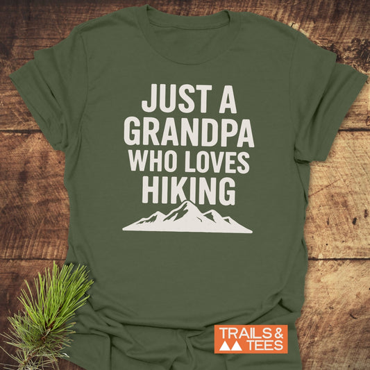 Hiking Grandpa T-Shirt with bold white text and mountain silhouette, laid flat on wood. Includes a rustic pine sprig and “TRAILS & TEES” logo.