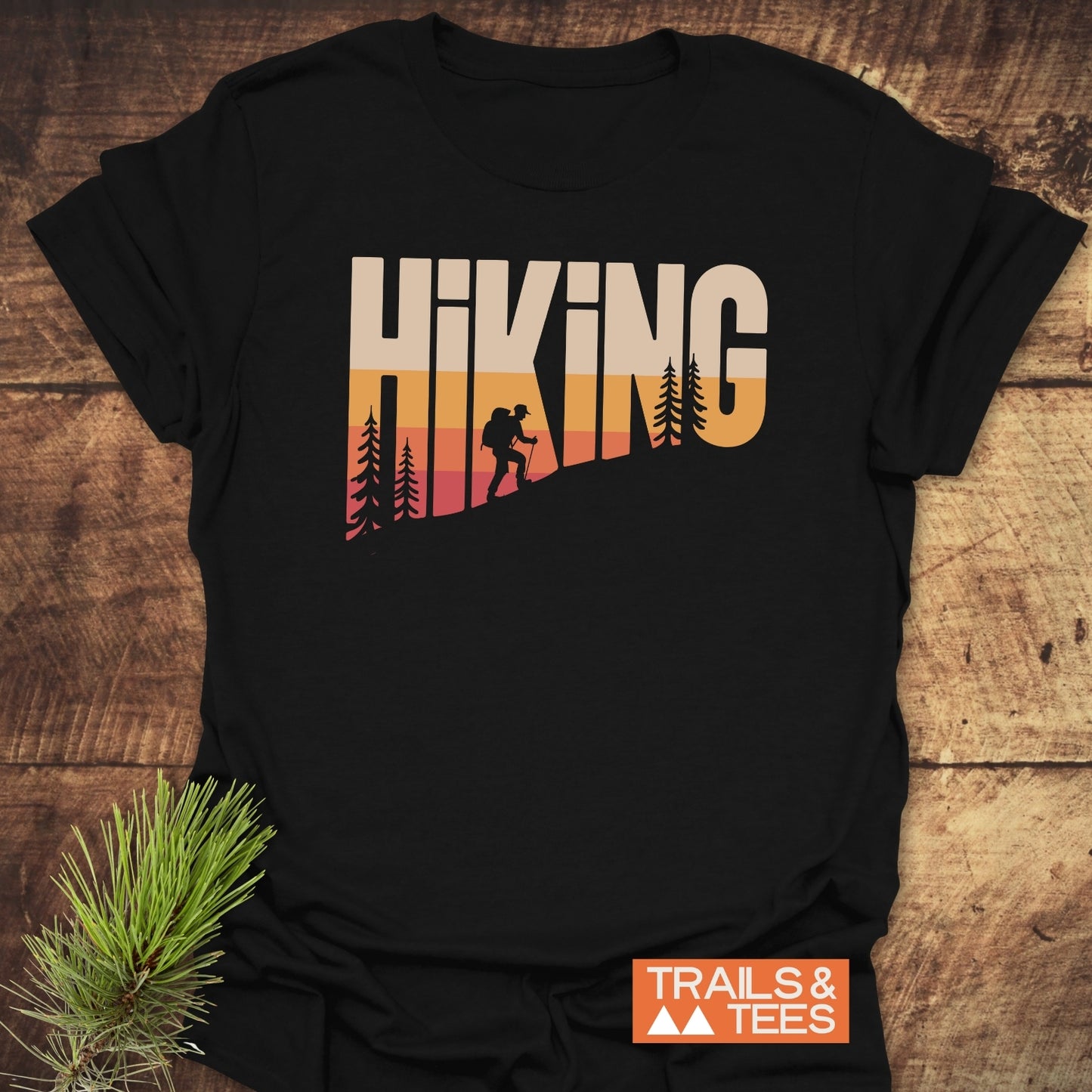 Hiking Vintage T-Shirt with sunset gradient HIKING graphic and hiker silhouette, laid on wood. Includes TRAILS & TEES logo and pine branch accent.