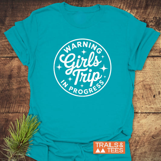 Girls Trip Hiking T-Shirt on wooden surface, featuring a WARNING Girls Trip IN PROGRESS badge. Crafted from soft, lightweight cotton with durable print, ideal for outdoor adventures.