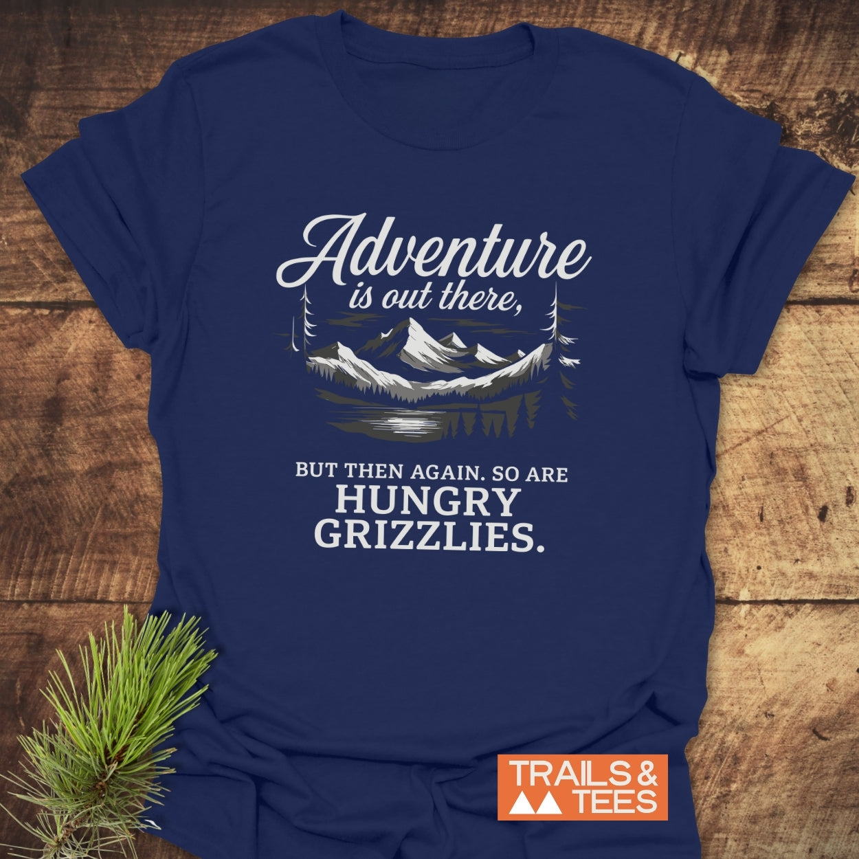 Navy-blue Adventure Is Out There T-Shirt with mountain illustration and humorous text, featuring an orange Trails & Tees logo, placed on a wooden surface with pine sprig.