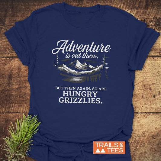 Navy-blue Adventure Is Out There T-Shirt with mountain illustration and humorous text, featuring an orange Trails & Tees logo, placed on a wooden surface with pine sprig.