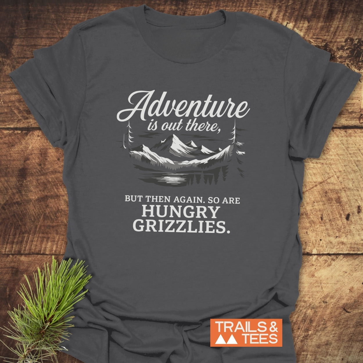 Adventure Is Out There T-Shirt with mountain graphic on a rustic wood surface, showcasing durable, soft cotton design by Trails & Tees, perfect for outdoor lovers.