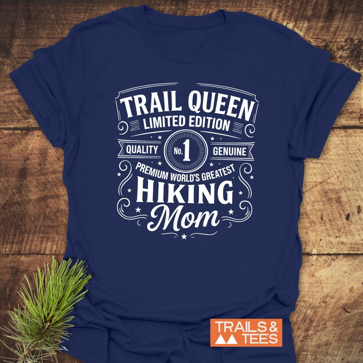 Trail Queen Mom T-Shirt with white typography, laid on wood, featuring a pine branch and Trails & Tees logo, emphasizing comfort and adventure.