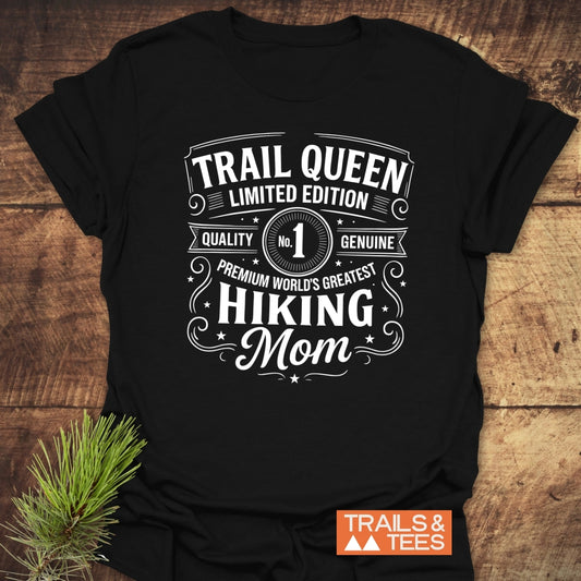 Trail Queen Mom T-Shirt with vintage-style white lettering on black fabric, laid on rustic wood; features TRAILS & TEES badge and pine sprig accent.