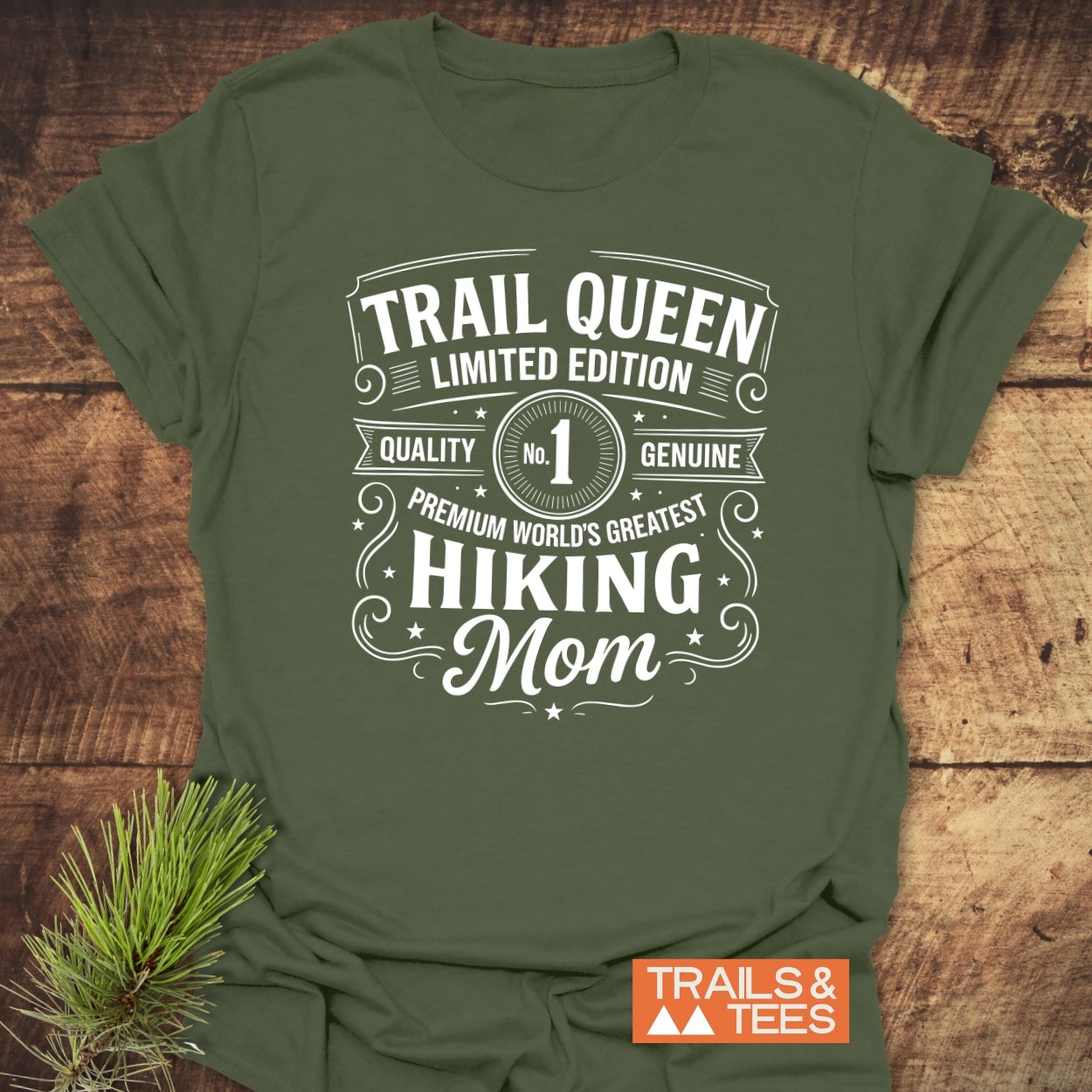 Forest-green Trail Queen Mom T-Shirt with white lettering, laid on wood; features premium quality, hiking mom design. Includes Trails & Tees logo and pine sprig.