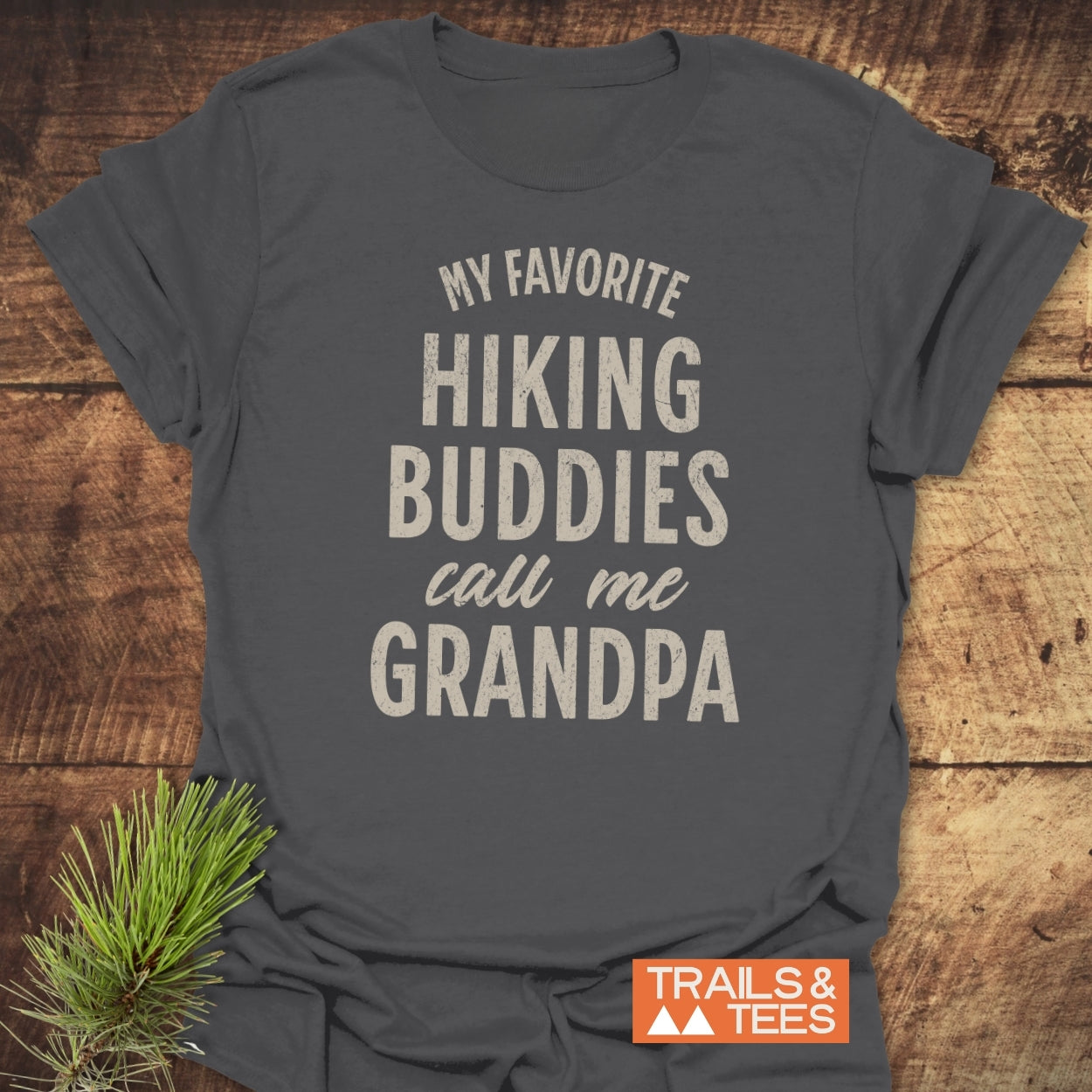 Dark-gray Hiking Grandpa T-Shirt with cream text. Laid flat on wood, features My favorite HIKING BUDDIES call me GRANDPA. Pine branch nearby.