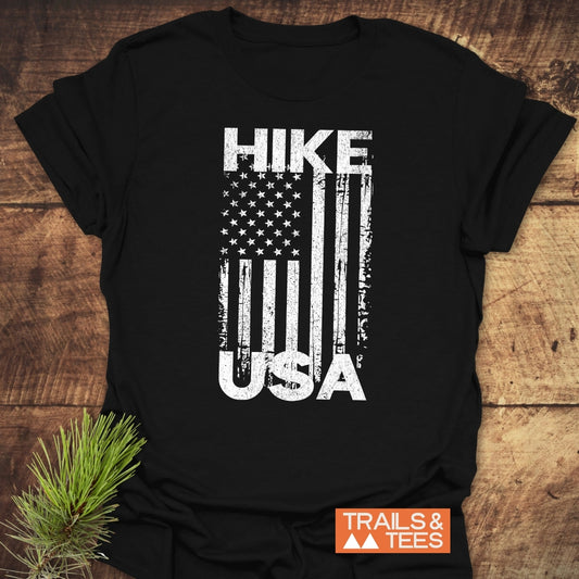 Hike USA T-Shirt featuring a distressed U.S. flag graphic on soft, lightweight cotton, laid on wood with a pine sprig, Trails & Tees logo visible.