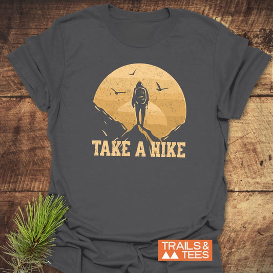 Take A Hike T-Shirt featuring a lone hiker graphic on a mountaintop, crafted from soft ring-spun cotton, laid flat with pine sprig beside it.