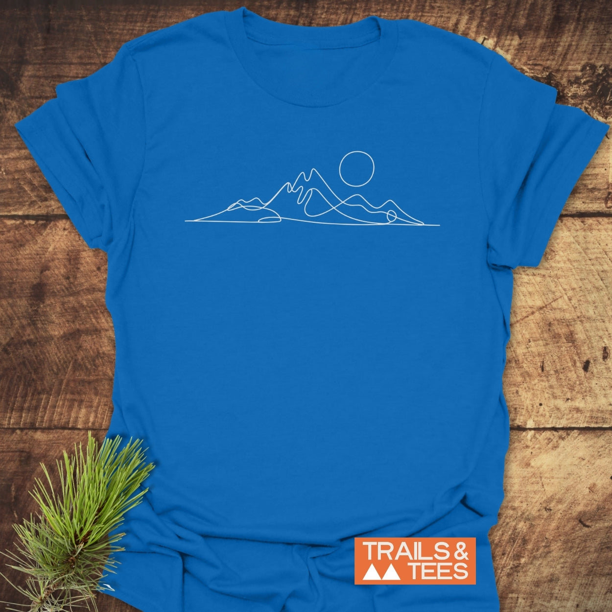Minimalist Mountains T-Shirt featuring a white mountain and sun outline, orange “TRAILS & TEES” logo, laid on wood with pine sprig for outdoorsy appeal.