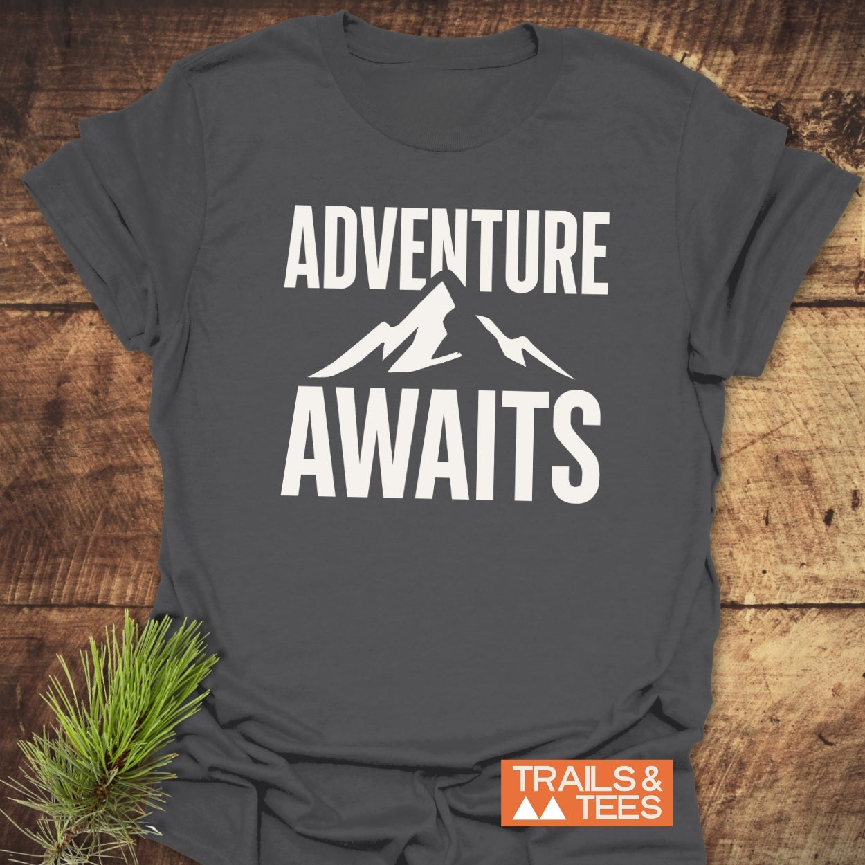 Adventure Awaits T-Shirt featuring bold ADVENTURE AWAITS text and mountain design, crafted from soft ring-spun cotton, laid flat with Trails & Tees logo.