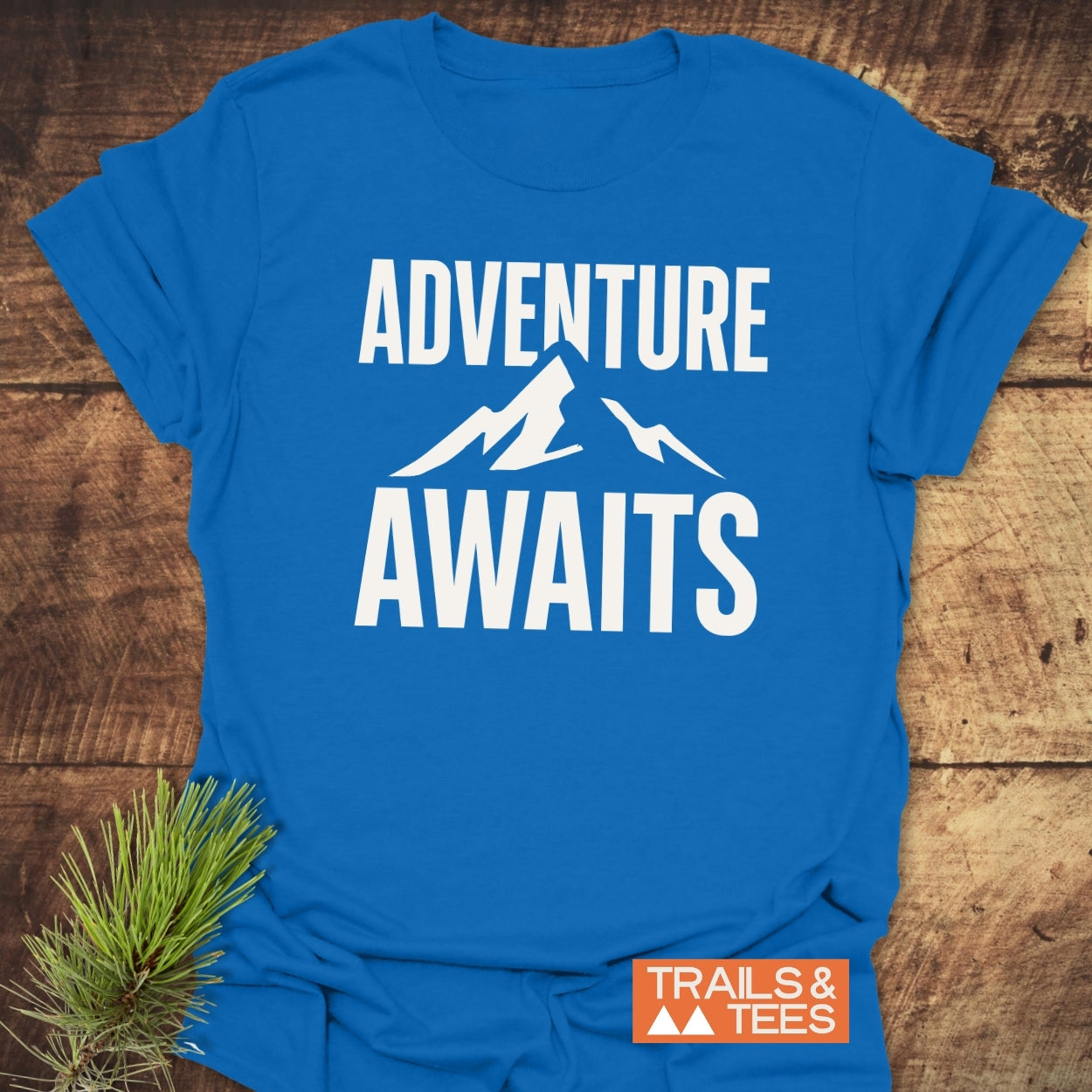 Adventure Awaits T-Shirt with bold white lettering and mountain silhouette, crafted from soft cotton, showcasing durability and outdoor spirit with Trails & Tees logo.