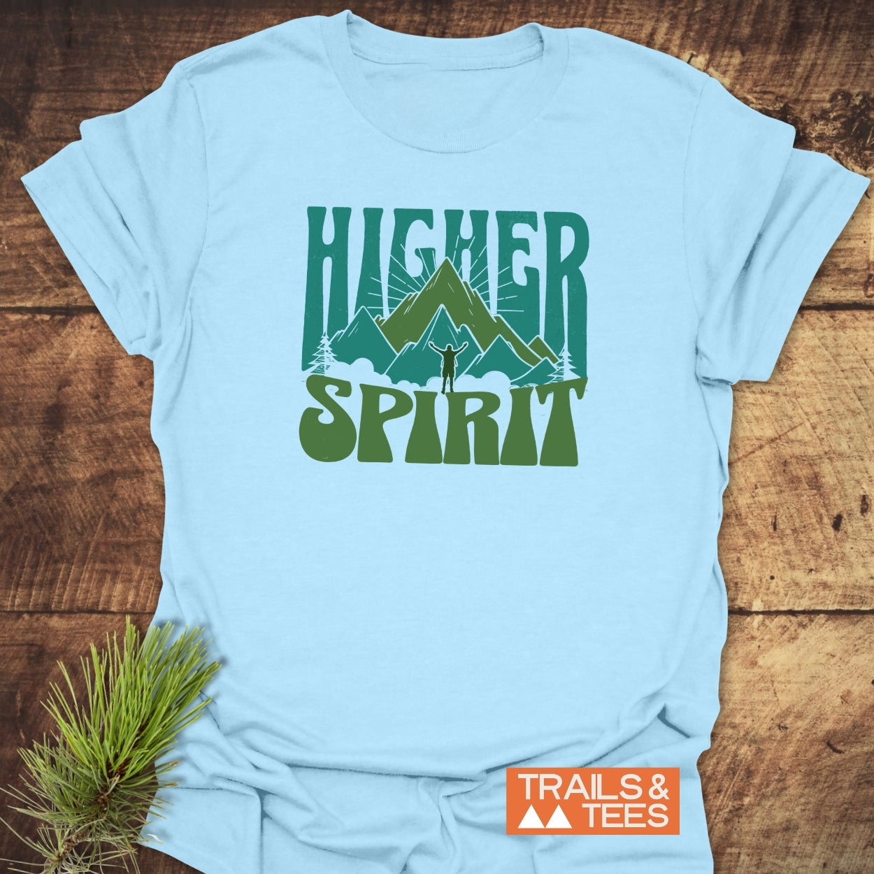 Higher Spirit T-Shirt featuring green mountain graphic and hiker silhouette on soft, light-blue fabric, perfect for outdoor enthusiasts. Includes TRAILS & TEES logo.