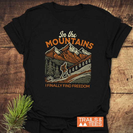 Mountain Freedom T-Shirt featuring a retro hiker graphic with pine trees and mountains, promoting outdoor adventure and freedom, laid flat on a wooden surface.