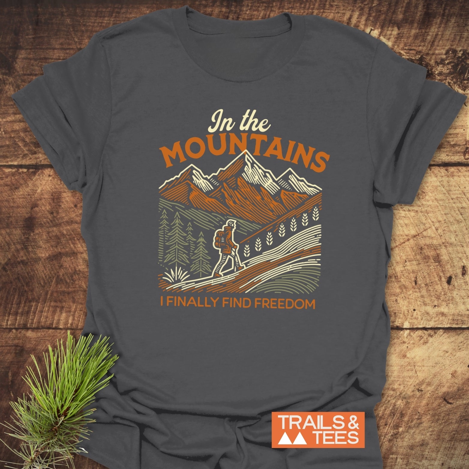 Mountain Freedom T-Shirt on wooden surface, features retro mountain scene with backpacker. Text reads “In the MOUNTAINS, I FINALLY FIND FREEDOM.” Includes Trails & Tees logo.
