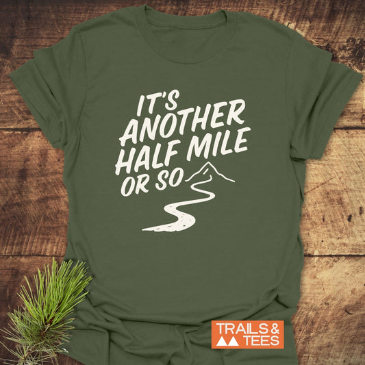 Half Mile Funny T-Shirt on a wooden surface, featuring a trail and mountain graphic. Showcases comfort, durability, and outdoor spirit with TRAILS & TEES logo.