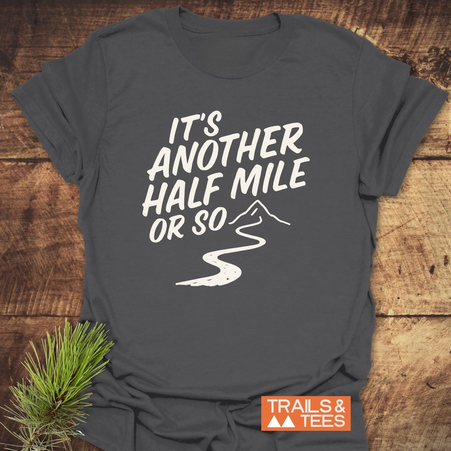 Half Mile Funny T-Shirt with slogan and mountain graphic, laid on wood surface. Features soft cotton, durable print, and rustic pine sprig accent.