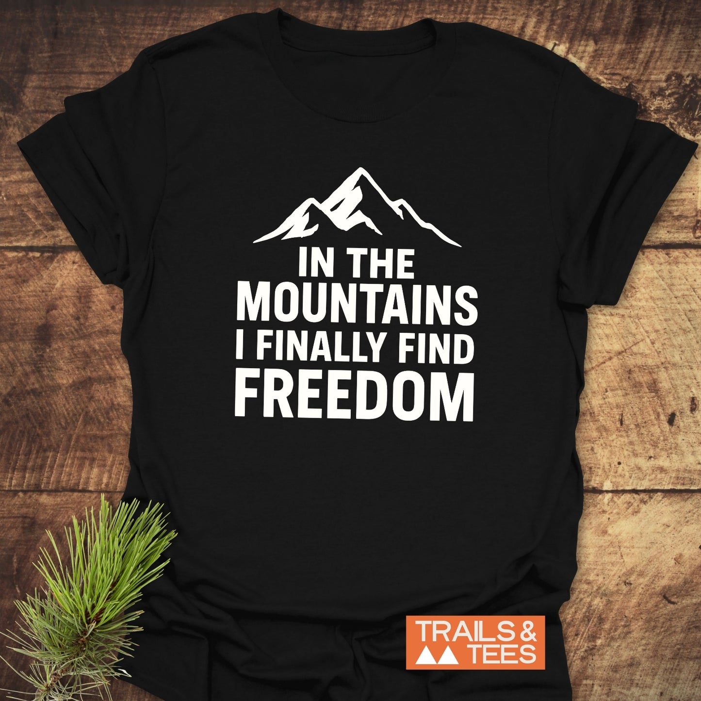 Finally Freedom T-Shirt on wooden surface, features a mountain graphic and bold text about freedom. Includes TRAILS & TEES logo. Made from soft, durable cotton.