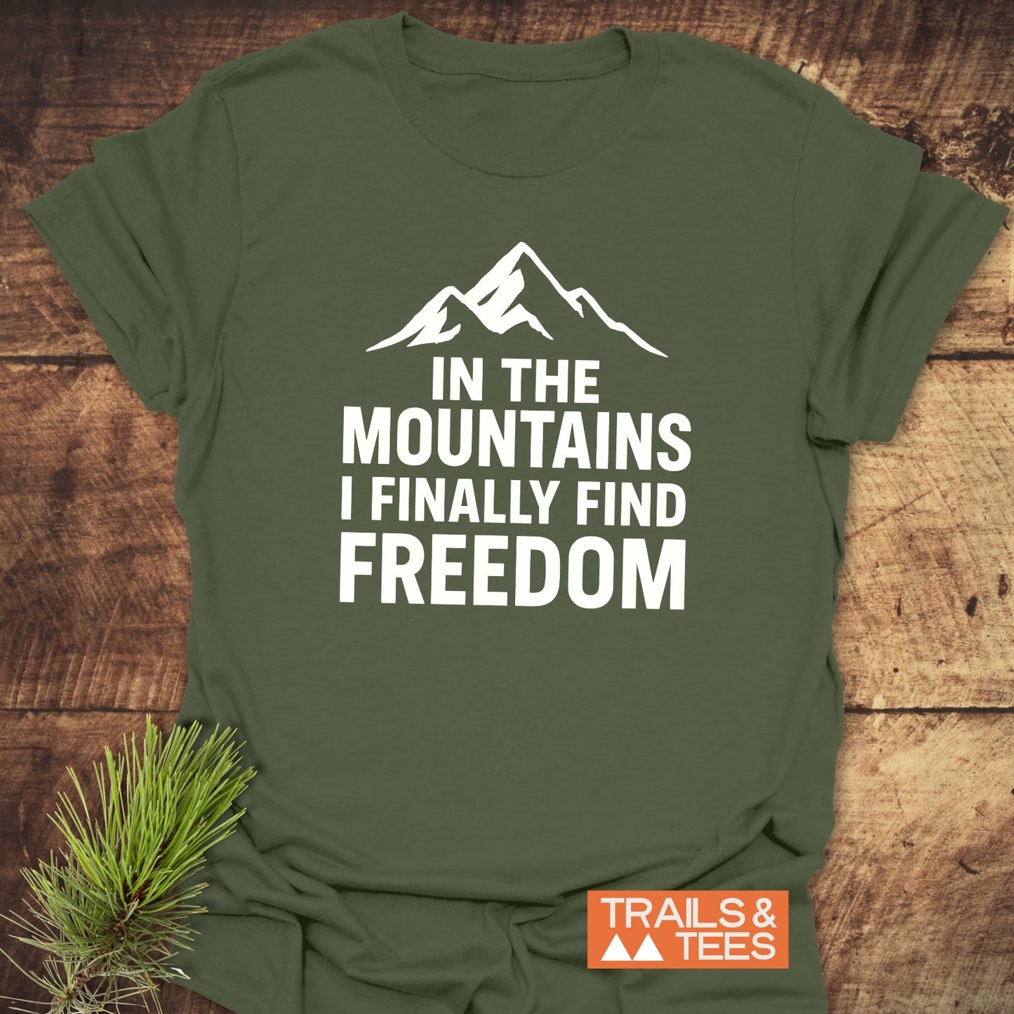 Finally Freedom T-Shirt showcasing a mountain silhouette and quote on olive fabric, highlighting its comfort and durability with a Trails & Tees logo.