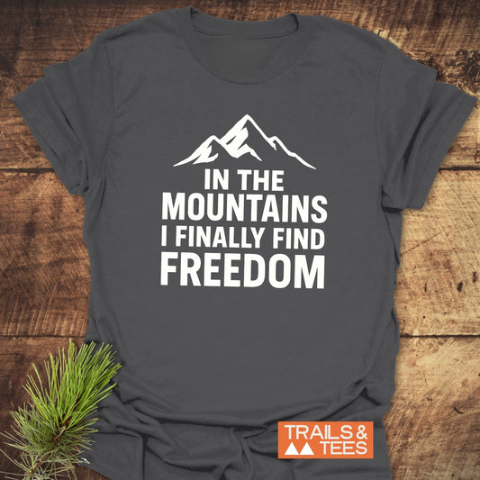 Charcoal-gray Finally Freedom T-Shirt with mountain silhouette and quote, laid on wood with pine sprig. Features soft cotton, durable print, pre-shrunk fit.