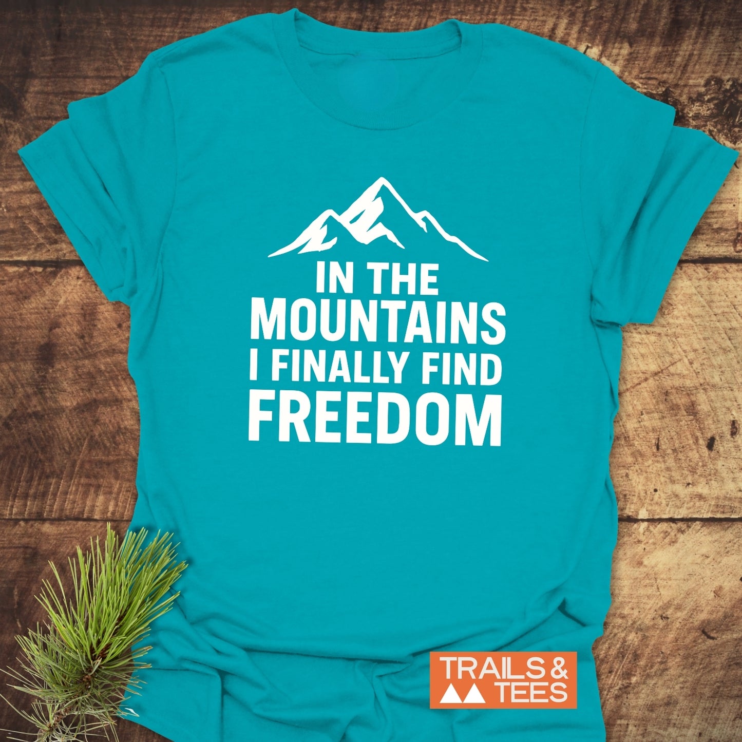 Finally Freedom T-Shirt with a mountain silhouette and motivational text in white, placed on rustic wood with a pine sprig, showcasing adventure-ready design.
