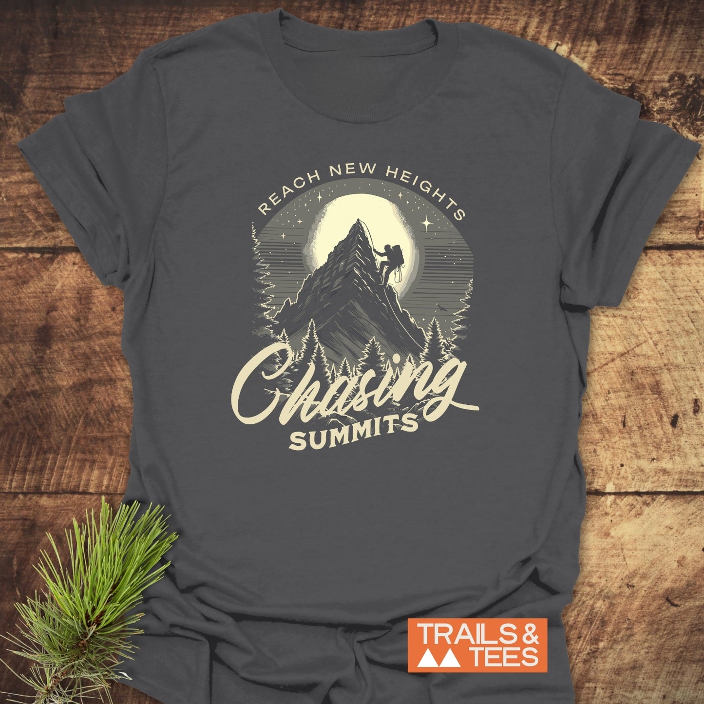 Chasing Summits T-Shirt featuring a night mountain climb design with “REACH NEW HEIGHTS” text, laid on wood with a pine sprig, logo nearby.