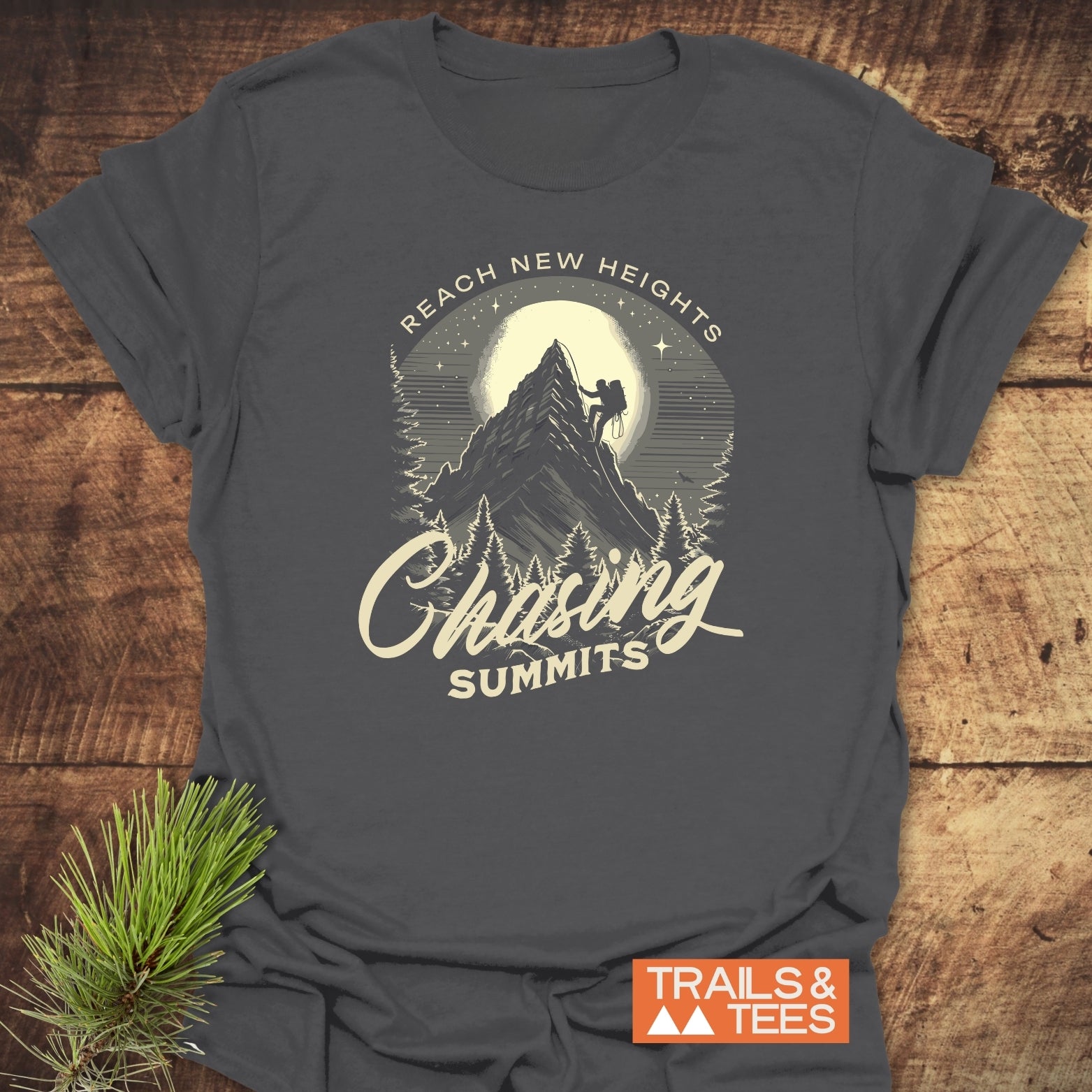 Chasing Summits T-Shirt featuring a night mountain climb design with “REACH NEW HEIGHTS” text, laid on wood with a pine sprig, logo nearby.