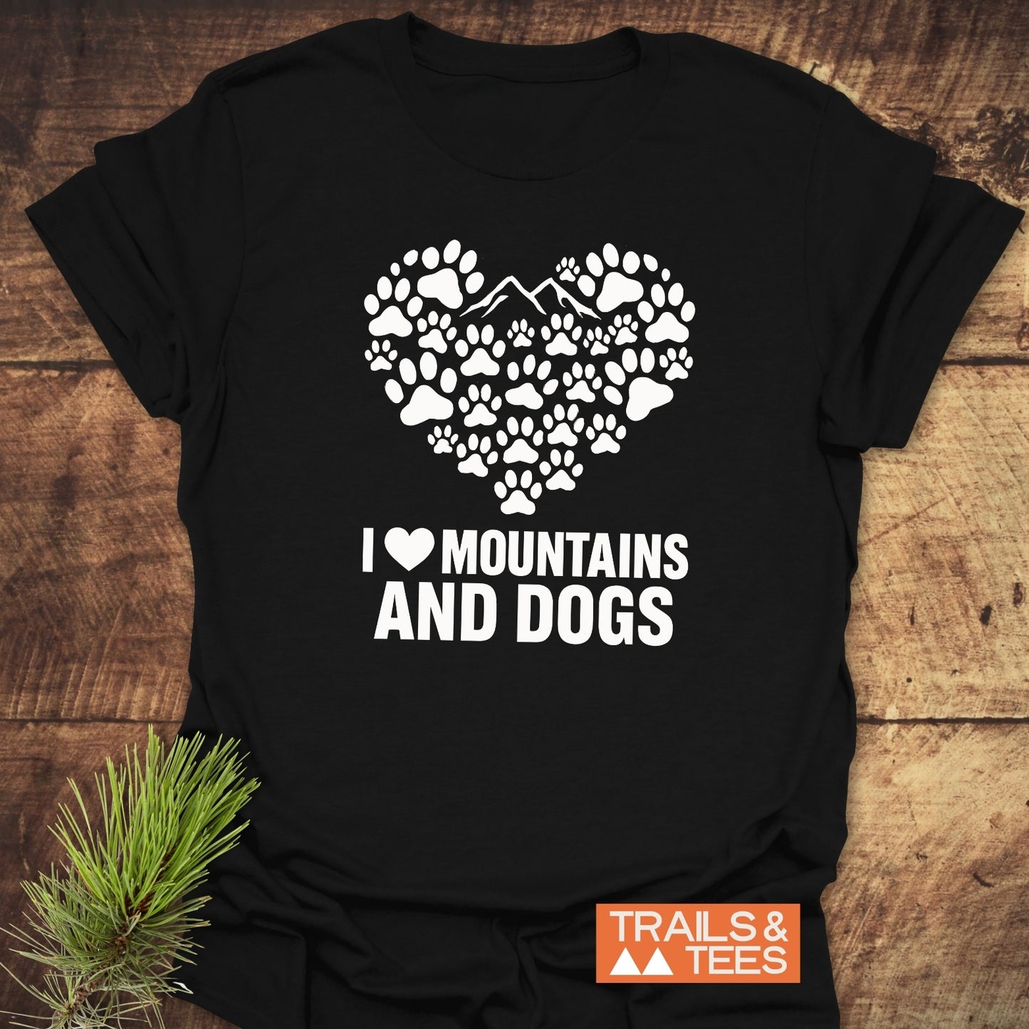 Paw Heart Dog Lover T-Shirt featuring white paw-print heart with mountain peaks, slogan “I ♥ MOUNTAINS AND DOGS,” laid on wooden surface with pine sprig.