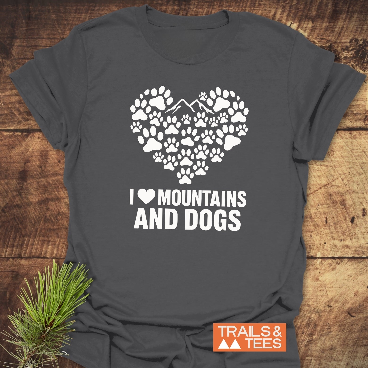 Charcoal gray Paw Heart Dog Lover T-Shirt with white paw and mountain heart design, slogan I ♥ MOUNTAINS AND DOGS, beside pine bough on wooden surface.