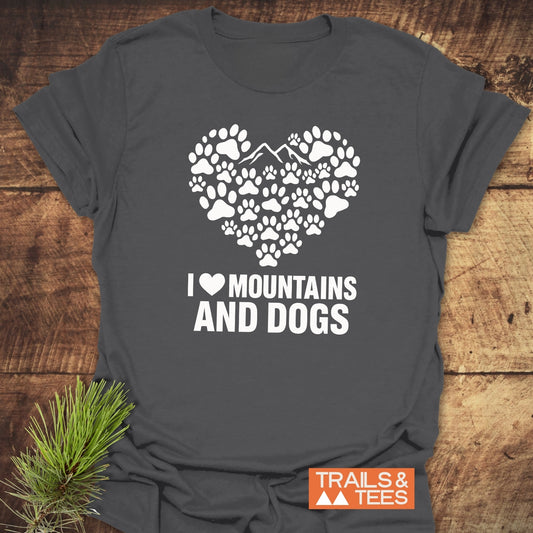 Charcoal gray Paw Heart Dog Lover T-Shirt with white paw and mountain heart design, slogan I ♥ MOUNTAINS AND DOGS, beside pine bough on wooden surface.